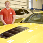 Male student wearing red shirt standing behind yellow Camaro.