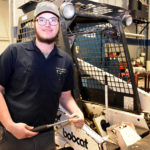 Male student holding wrench standing in front of bobcat vehicle.