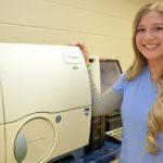 Female student in blue scrubs standing next to medical lab equipment.