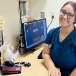 Female nurse wearing blue scrubs, smiling, sitting at a desk in a clinic room.